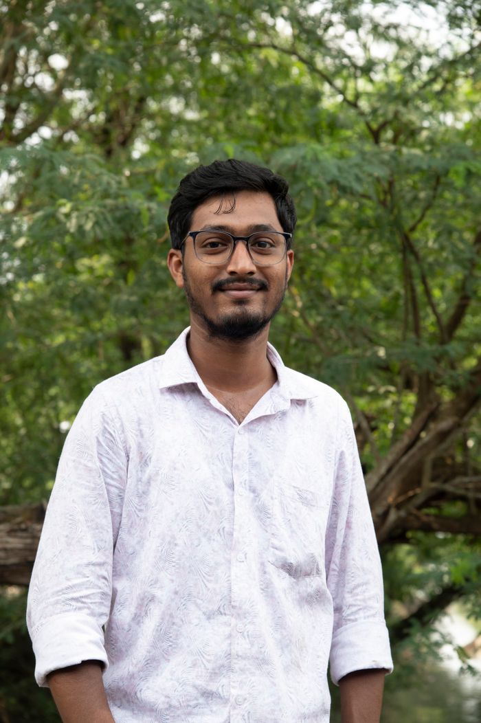 A young man smiling in a natural outdoor setting, surrounded by greenery.